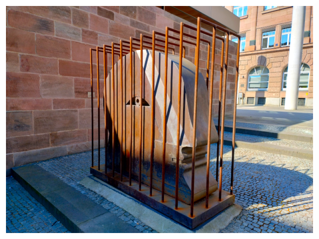 The image shows a large sculpture of a human face enclosed in a rectangular cage made of vertical metal bars. The face is stylized, with distinctive features such as the nose and mouth, and appears to be made of smooth material. The cage sits on a stone base set on a cobblestone surface. The sculpture is located near a brick wall, and in the background is a building with large windows. The cage's bars are evenly spaced and extend above the height of the sculpture.