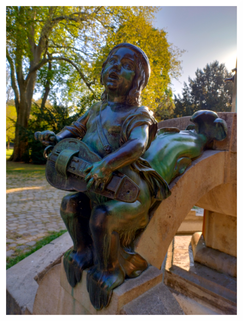 The image shows a bronze statue of a child sitting on a stone structure. The child holds a stringed instrument, possibly a guitar or lute, in both hands. The statue is detailed and shows the child's clothing and facial features. The background features trees with thick foliage, suggesting an outdoor atmosphere. The statue stands on a stone base and the surrounding area appears to be paved.